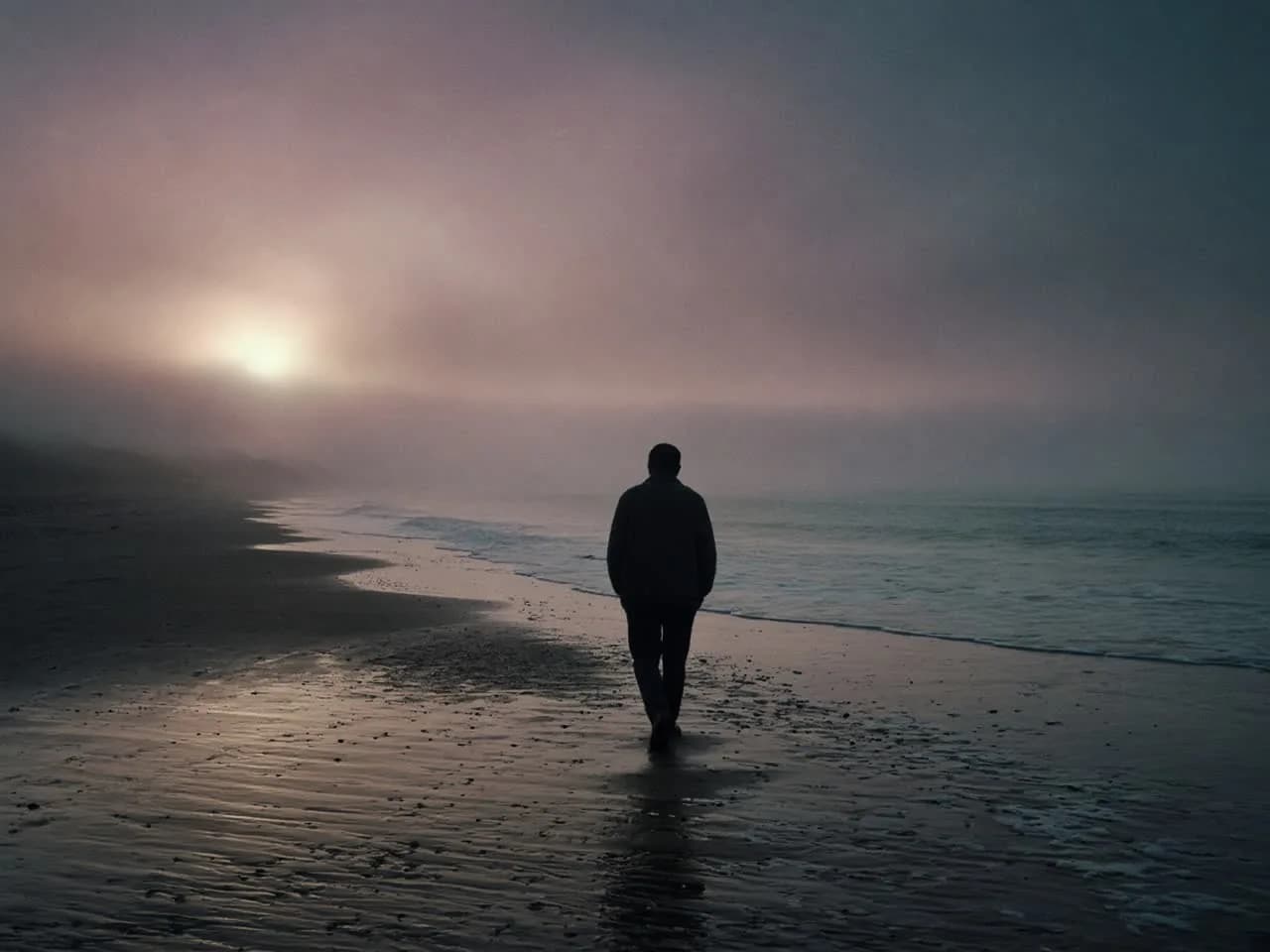 A person in a dark sweater walking a wet crescent of packed sand at dusk, back to camera, a pale sun low left.